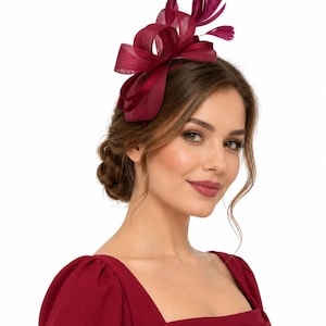 May include: A deep burgundy fascinator headpiece featuring a structured, looped sinamay bow design accented with delicate, upright feathers. The headpiece is secured on a thin, matching headband, creating a cohesive and elegant hair accessory.