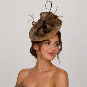May include: A dark brown fascinator hat featuring a circular base with a textured, woven finish. The hat is adorned with a large, structured bow made of matching brown fabric, delicate upright feathers, and a thin, curved wire accent.