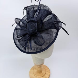 Op de afbeelding: Een marineblauwe fascinator hoed met een cirkelvormige basis en een decoratieve strik. De hoed heeft een geweven textuur en is versierd met veren en een gekruld accent. Het wordt getoond op een mannequinhoofd.