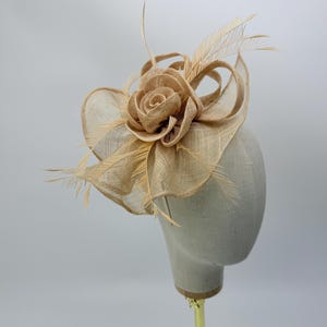 Op de afbeelding: Een beige fascinator met een stoffen roos en delicate veren. De hoofdbedekking wordt getoond op een mannequinhoofd. De roos is het middelpunt, omringd door geluste stof en ijle veren, wat een elegant ontwerp creëert.