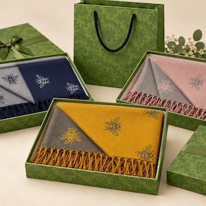 May include: Three rectangular gift boxes containing folded scarves with bee patterns. One box holds a navy and grey scarf, another a pink and grey scarf, and the third a mustard yellow and grey scarf. Each scarf features fringe and matching gift boxes.