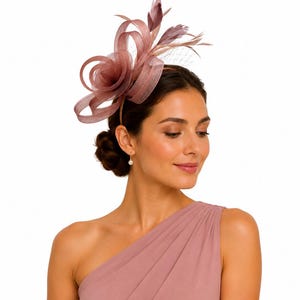 May include: A dusty rose fascinator headpiece featuring structured sinamay loops, delicate feathers, and a fine netting veil. The headpiece is mounted on a thin headband, showcasing a monochromatic design with layered textures and a sculptural form.