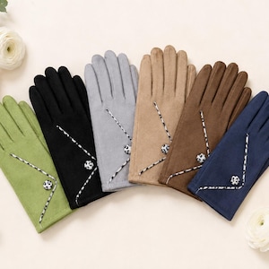 Könnte beinhalten: Eine Kollektion von sechs Handschuhen in Grün, Schwarz, Grau, Beige, Braun und Marineblau. Jeder Handschuh verfügt über ein diagonal genähtes Detail mit einer schwarz-weiß gemusterten Kordel und einem passenden runden Knopf auf dem Handrücken.