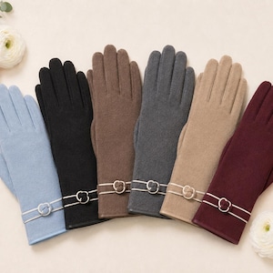 May include: A collection of six pairs of gloves in light blue, black, brown, dark grey, beige, and burgundy. Each pair features a smooth, solid-coloured fabric with a decorative double-strap detail at the wrist, accented by a small metal ring.