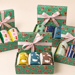 May include: Four rectangular green gift boxes with a pink floral pattern and a light pink satin ribbon bow. Each box contains three pairs of rolled socks featuring various patterns including cherries, bees, and small animals on colourful backgrounds.
