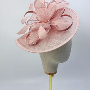 Może przedstawiać: Pudrowo-różowy kapelusz typu fascinator z szeroką, okrągłą podstawą wykonaną z teksturowanej, tkanej tkaniny. Wierzch ozdobiony jest misternymi pętlami z pasującej półprzezroczystej wstążki oraz kilkoma delikatnymi, miękkimi piórami w kwiatowym układzie.