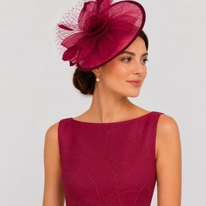 Pode incluir: Um fascinator magenta profundo com base circular, uma flor de tecido central e rede delicada. O chapéu é combinado com um vestido sem mangas a condizer que apresenta um padrão subtil de círculos cosidos tom sobre tom no tecido.