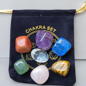 Op de afbeelding: Een Chakra Set gepresenteerd in een zwarte fluwelen zakje. Het zakje heeft gouden tekst met de tekst "CHAKRA SET". De set bevat zeven gepolijste edelstenen in verschillende kleuren: oranje, paars, helder, groen, roze, blauw en geelbruin.