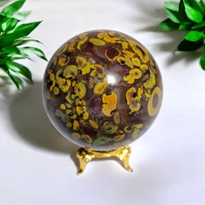 Puede incluir: Una piedra esférica pulida con una base de color marrón oscuro y patrones circulares e irregulares en tonos amarillo, dorado y rojo intenso. La esfera descansa sobre un pequeño soporte metálico dorado de tres patas con acabado liso.