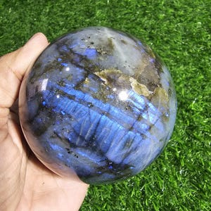 Op de afbeelding: Een gepolijste labradoriet bol met een glad, rond oppervlak met diepblauwe iriserende flitsen, donkergrijze insluitingen en doorschijnende plekken. De steen vertoont natuurlijke minerale aders en een reflecterende, glanzende afwerking.