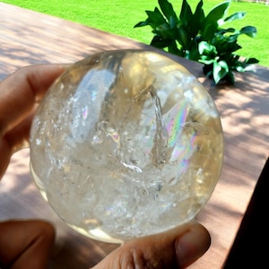 Könnte beinhalten: Eine klare, kugelförmige Kristallkugel mit inneren Brüchen und Regenbogenreflexionen. Der Kristall wird in einer Hand gehalten, mit einer Holzoberfläche und Grün im Hintergrund. Die Kugel hat einen gelblichen Farbton.