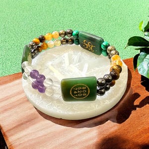 Puede incluir: Una pulsera de cuentas con piedras preciosas de colores y detalles de jade verde. La pulsera incluye una variedad de cuentas en tonos amarillos, marrones, verdes y morados. Dos rectángulos de jade verde están grabados con símbolos.