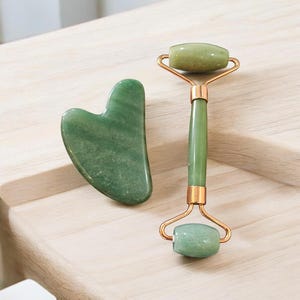 May include: A green jade facial roller and a heart-shaped gua sha tool. The roller has a gold-colored frame and handle. The tools are displayed on a light-colored wooden surface.