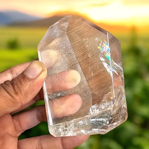 Puede incluir: Una punta de cristal de cuarzo transparente y pulido con forma hexagonal y base plana. El cristal presenta fracturas internas, inclusiones naturales y una refracción de luz de colores del arcoíris en una de sus facetas anguladas.