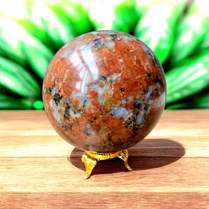 Pode incluir: Uma pedra polida de forma esférica com um padrão manchado de minerais em tons de laranja profundo, vermelho ferrugem, cinzento e preto. A esfera repousa sobre um pequeno suporte metálico dourado de três pernas com pés curvos e ornamentados.