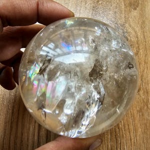 Puede incluir: Una esfera de cristal de cuarzo transparente con fracturas internas e inclusiones naturales. La superficie pulida refleja la luz, creando refracciones prismáticas de colores del arcoíris dentro de la estructura de la piedra.