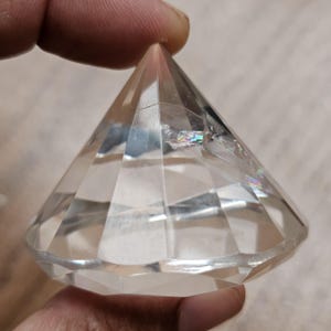 Può includere: Un cristallo trasparente sfaccettato a forma di diamante con una punta superiore e una base ampia a molteplici facce. Il materiale trasparente presenta fratture interne che creano piccoli riflessi di luce arcobaleno all'interno.