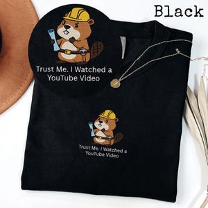May include: Black t-shirt featuring an embroidered cartoon beaver wearing a yellow hard hat and holding blueprints. The text "Trust Me. I Watched a YouTube Video" is below the beaver. A gold necklace is draped on the shirt.