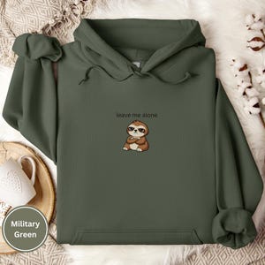 May include: Military green hoodie with the text "leave me alone" and a cartoon sloth with crossed arms. The hoodie has a drawstring hood and a front pocket. The background is a soft, neutral color.
