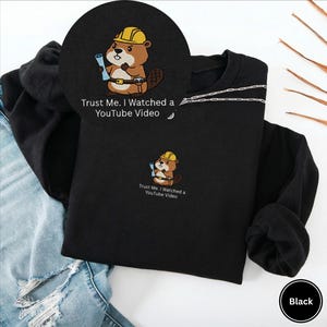 May include: Black sweatshirt featuring an embroidered cartoon beaver wearing a yellow hard hat and holding blueprints. The text "Trust Me. I Watched a YouTube Video" is below the beaver. The sweatshirt is shown on a white background.