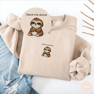 May include: Sand-colored sweatshirt featuring a cartoon sloth with crossed arms and the phrase "leave me alone." The sloth is brown and beige, with a grumpy expression. A small silver necklace is visible. The sweatshirt is folded.