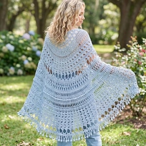 Può includere: Uno scialle all'uncinetto azzurro chiaro con un motivo traforato e un bordo smerlato con piccole nappe. Lo scialle è drappeggiato sulle spalle. Lo sfondo mostra un giardino con alberi e vegetazione.