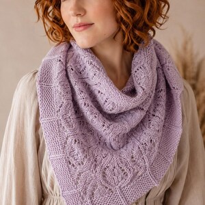 May include: A lavender-colored knitted shawl with a detailed openwork pattern. The shawl is draped around the shoulders, showcasing the intricate design and soft texture. The knitted design features geometric and leaf-like shapes.
