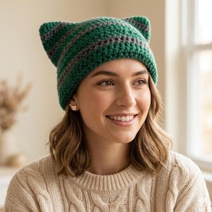 May include: A green and grey striped crochet cat ear hat. The handmade hat has a ribbed texture. The hat is worn by a person wearing a cream-coloured sweater.