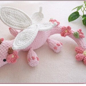 May include: A handmade pink crochet dragon with white wings and floral accents. The dragon has a textured body, small black eyes, and is decorated with pink and brown flowers. The wings are white with a textured border.