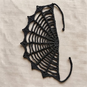 May include: A black crochet piece shaped like a spider web with a semi-circular design. The item features a series of radiating spokes connected by concentric arched rows, ending in pointed edges and two long, thin crocheted ties.