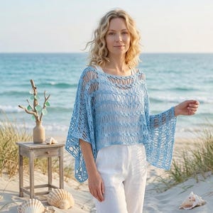 Può includere: Un top all'uncinetto azzurro con maniche a campana. Il top è indossato sopra una canotta bianca e abbinato a pantaloni bianchi. L'immagine è scattata su una spiaggia sabbiosa con l'oceano sullo sfondo.
