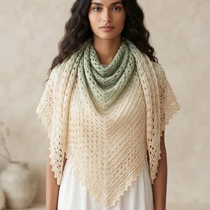May include: A triangular crochet shawl featuring an ombre color transition from a soft sage green at the neckline to a creamy off-white at the pointed hem. The garment displays an intricate openwork stitch pattern with a scalloped decorative edge.