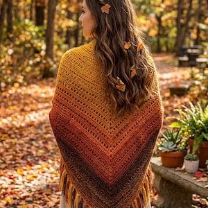 May include: A handmade, crochet shawl in a gradient of autumn colors: yellow, orange, and brown. The shawl has a triangular shape with a fringed edge. The person is standing in a forest with fallen leaves.