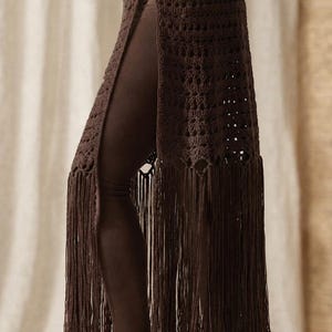 Puede incluir: Una falda de ganchillo marrón oscuro con una abertura lateral, dos botones dorados en la cintura alta y largos flecos en el dobladillo inferior. La prenda se lleva sobre unos leggings marrón oscuro, mostrando un patrón de punto calado.