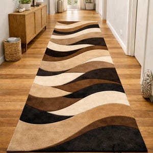 Könnte beinhalten: Ein langer, rechteckiger Läufer mit einem sich wiederholenden Wellenmuster in den Farben Creme, Beige, Mittelbraun und Dunkelanthrazit. Der Teppich hat eine strukturierte, geschnittene Floroberfläche und enthält den Text City Carpets In.