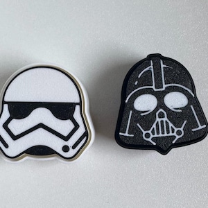 Può includere: Due toppe a tema personaggi di Star Wars. Una è un casco da Stormtrooper bianco con dettagli neri e un contorno dorato. L'altra è un casco nero di Darth Vader con dettagli bianchi.