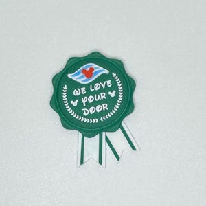 Peut inclure: Un badge vert de style récompense circulaire avec le texte blanc "We Love Your Door". Le badge présente une tête de Mickey Mouse et un graphique de navire. Deux rubans blancs et verts pendent en dessous.