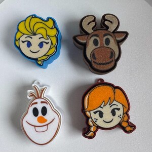 Könnte beinhalten: Vier farbenfrohe Charakteranhänger mit Elsa mit gelbem Haar, einem braunen Rentier, Olaf dem Schneemann und Anna mit orangefarbenen Zöpfen. Jeder Anhänger hat eine Schlaufe zum Befestigen an einem Armband oder einer Halskette.