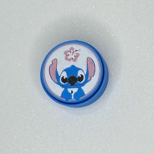 May include: A blue, circular button featuring a cartoon character with blue fur, large ears, and a black nose. The character is set against a white background with a pink flower. The button has a blue border.