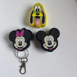 May include: Three cartoon character keyrings: Mickey Mouse, Minnie Mouse, and Pluto. Mickey and Minnie are black with white faces and pink mouths. Minnie has a pink bow. Pluto is yellow with a black outline and a pink tongue.