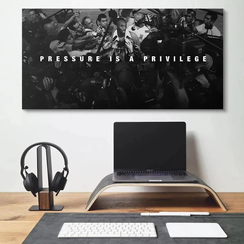 Op de afbeelding: Een zwart-wit wandprint met een menigte mensen rondom een centrale figuur met de tekst PRESSURE IS A PRIVILEGE in het midden in witte hoofdletters. Het kunstwerk hangt boven een bureau met een laptop en een koptelefoon.