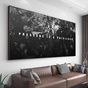 Op de afbeelding: Een grote rechthoekige zwart-wit wandprint met een menigte fotografen met camera's rondom een centrale figuur. De tekst "PRESSURE IS A PRIVILEGE" is in dikke witte hoofdletters in het midden van de afbeelding gedrukt.