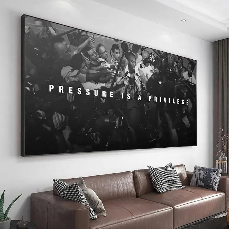 Op de afbeelding: Een grote rechthoekige zwart-wit wandprint met een menigte fotografen met camera's rondom een centrale figuur. De tekst "PRESSURE IS A PRIVILEGE" is in dikke witte hoofdletters in het midden van de afbeelding gedrukt.