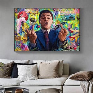 Può includere: Una stampa artistica da parete rettangolare che raffigura il ritratto di un uomo in abito blu e cravatta, che fa un gesto con entrambi i medi alzati. Lo sfondo è un vivace collage di graffiti multicolore con varie forme, simboli e testi astratti.