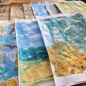 Könnte beinhalten: Eine Sammlung von Aquarellmalereien, die Küstenszenen darstellen. Die Kunstwerke zeigen blaue, grüne und gelbe Farbtöne mit Bildern von Wellen, Möwen und Sandstränden. Die Gemälde sind auf weißem Papier.