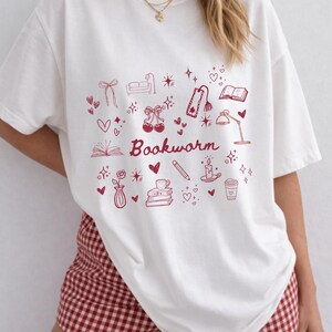 Könnte beinhalten: Weißes T-Shirt mit rotem Grafikdesign, das das Wort "Bookworm" und verschiedene Illustrationen zeigt, darunter Bücher, Herzen und eine Kaffeetasse. Das Shirt wird mit rot-weiß karierten Shorts getragen.