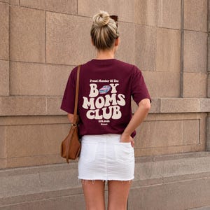 May include: A maroon t-shirt with the text "Proud Member Of The Boy Moms Club" in white, with a car graphic. The shirt is paired with a white denim skirt. A woman is wearing the shirt, with a brown purse and sunglasses.