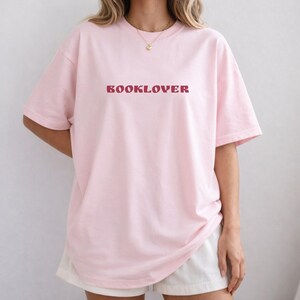 May include: A light pink t-shirt with the word "BOOKLOVER" printed in a burgundy color across the chest. The shirt has short sleeves and a relaxed fit. The model is wearing white shorts and a gold necklace.