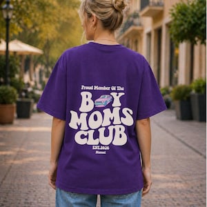 May include: A purple t-shirt with the text "Proud Member Of The Boy Moms Club" in white, along with a car graphic. The shirt also includes "EST.2026" and the name "Manuel" in a smaller font. The person is wearing blue jeans.