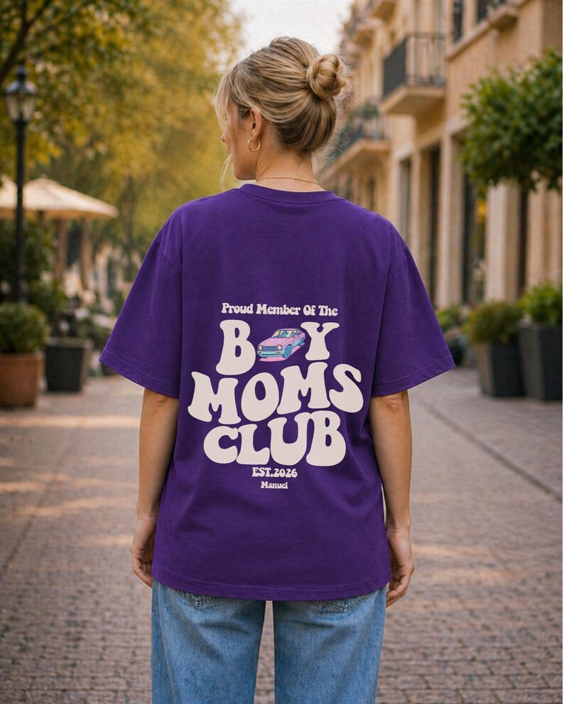 May include: A purple t-shirt with the text "Proud Member Of The Boy Moms Club" in white, along with a car graphic. The shirt also includes "EST.2026" and the name "Manuel" in a smaller font. The person is wearing blue jeans.